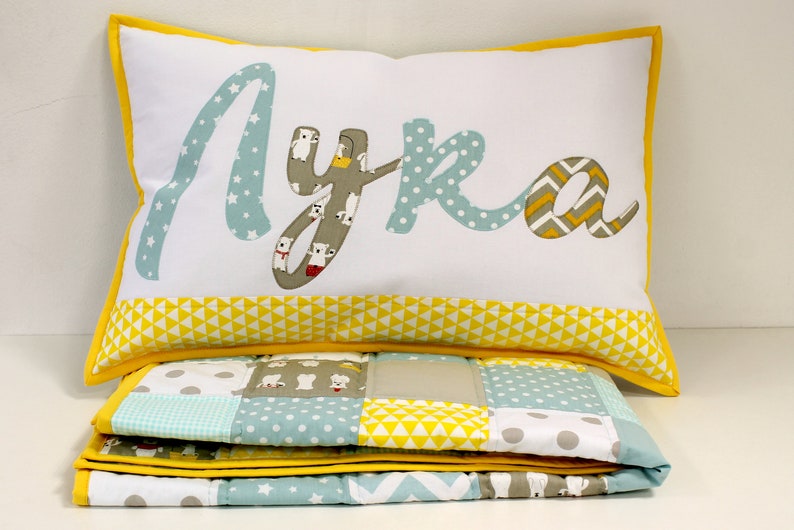 May include: A white pillow with a yellow border and the name "Lyra" spelled out in colorful fabric letters. The pillow is sitting on top of a folded quilt with a patchwork design in shades of blue, yellow, and gray.