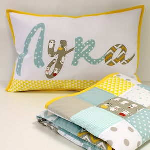 May include: A white pillow with a yellow border and the name "Lyka" spelled out in blue fabric with a white polka dot pattern. The pillow is sitting on top of a folded blue and white patchwork quilt with a yellow border.