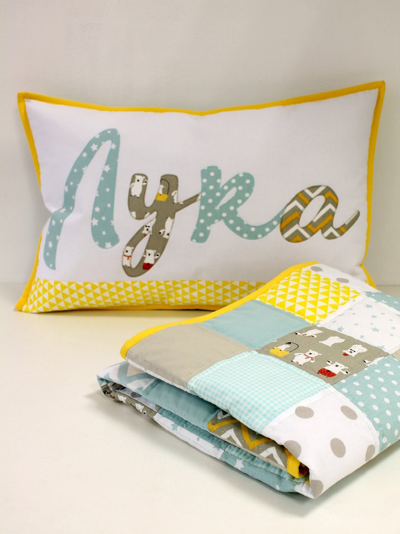 May include: A white pillow with a yellow border and the name "Lyka" spelled out in blue fabric with a white polka dot pattern. The pillow is sitting on top of a folded blue and white patchwork quilt with a yellow border.