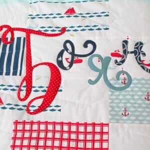 May include: A red, white and blue baby quilt with the name "Jack" spelled out in large, colourful letters. The quilt features a nautical theme with anchors, whales and sailboats.