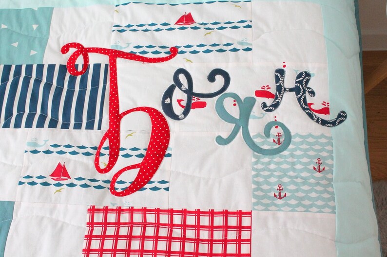 May include: A red, white and blue baby quilt with the name "Jack" spelled out in large, colourful letters. The quilt features a nautical theme with anchors, whales and sailboats.