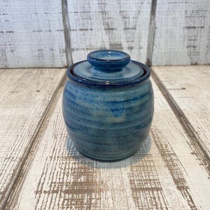 May include: A small, round, ceramic jar with a matching lid, featuring a variegated blue glaze with darker blue accents and horizontal ridges along the body. The jar has a smooth, glossy finish and a circular knob handle on the center of the lid.