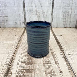 May include: A handmade ceramic tumbler with a textured, ridged exterior. The cup is a blend of blue and teal hues, with a darker blue interior. The cylindrical shape and rustic finish suggest a handcrafted aesthetic, suitable for beverages or as a decorative piece.