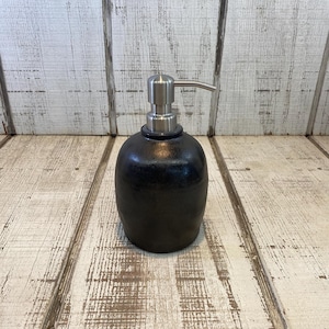 May include: A matte black soap dispenser with a rounded, bulbous body and a brushed silver metal pump mechanism. The container features a smooth, dark finish that contrasts with the metallic sheen of the top nozzle, designed for dispensing liquid soap.