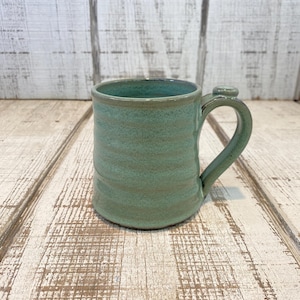 May include: A handmade ceramic mug in a seafoam green color. The mug has a rounded body with a textured surface and a curved handle. The interior and rim are a darker shade of green. The mug is approximately 4 inches tall.