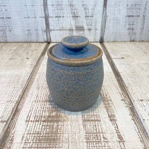 May include: A small, round, blue ceramic jar with a lid. The jar has a speckled texture and a light brown rim. The lid has a small knob on top. The jar is sitting on a light-colored wooden surface.