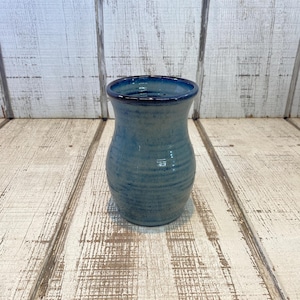 May include: A ceramic vase with a bulbous body and a flared rim, featuring a glossy blue glaze that transitions to a darker navy blue at the top edge. The surface displays subtle horizontal ridges and a smooth, reflective finish throughout.