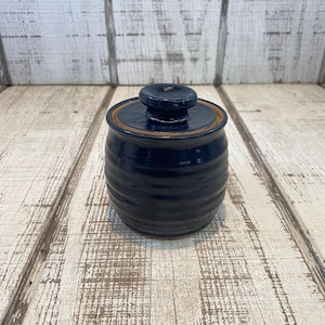 May include: A dark blue ceramic jar with a lid. The jar has a round shape with horizontal ridges and a brown rim. The lid is a matching dark blue color. The jar is sitting on a white and brown wooden surface.