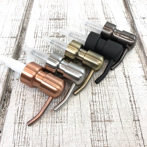May include: A collection of five metal bottle pour spouts in various finishes: copper, silver, gold, black, and gunmetal. Each spout has a clear plastic tube and a curved pouring spout. The spouts are arranged on a white wooden surface.