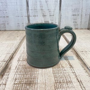 May include: A handmade ceramic mug in a teal green glaze. The mug has a rounded body, a curved handle, and a small decorative element where the handle meets the mug. The mug is sitting on a light-colored wooden surface.