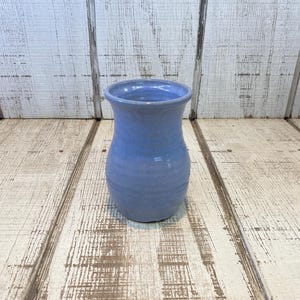 May include: A small, light blue ceramic vase with a smooth, glossy finish. The vase features a rounded, bulbous body that tapers slightly toward the neck before widening at the top rim, creating a simple, functional silhouette for holding flowers.