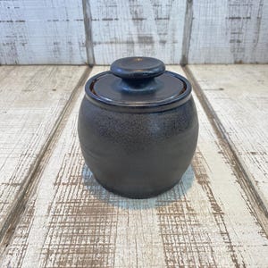 May include: A small, round, dark charcoal-colored ceramic jar with a matching lid. The jar features a smooth, slightly speckled matte finish and a central knob handle on the lid. The vessel has a bulbous body shape and a short, narrow neck.