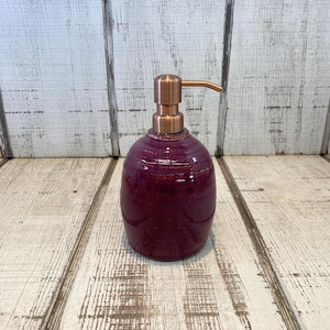 May include: A round, deep purple ceramic soap dispenser with a glossy, textured finish and a metallic copper-colored pump mechanism. The body of the dispenser features subtle variations in color intensity, creating a rich, multi-tonal appearance.