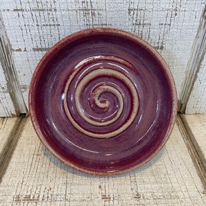 May include: A small, round, purple ceramic dish with a white swirl design in the center.