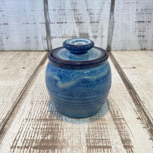 May include: A small, round ceramic jar with a lid, featuring a mottled blue glaze with darker blue and lighter cream-colored accents. The jar has a slightly tapered shape with subtle horizontal ridges along the lower body and a matching lid.
