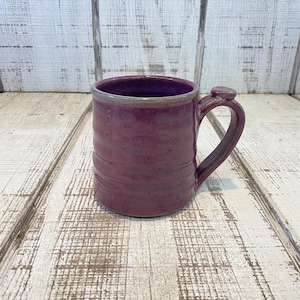 May include: A handmade ceramic mug in a deep plum color with a textured surface. The mug has a rounded handle and a small decorative detail where the handle meets the cup. The rim is a lighter shade of purple.