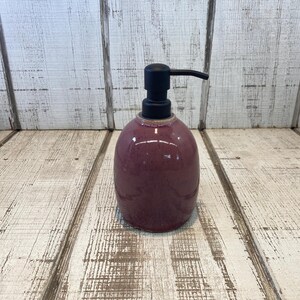 May include: A rounded ceramic soap dispenser with a glossy, speckled plum-colored finish and a matte black plastic pump mechanism. The container has a smooth, bulbous shape that tapers slightly toward the top, featuring a consistent dark purple glaze.