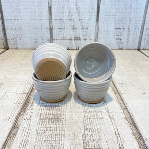 May include: A set of four small ceramic cups with a light gray glaze and horizontal ribbed texture. The cups feature a natural, unglazed brown clay base with a distinct wavy pattern on the bottom surface, showcasing a simple, functional design.