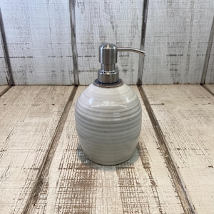 May include: A ceramic soap dispenser with a rounded, bulbous body featuring a light cream-colored glaze and horizontal ribbed texture. The top is fitted with a brushed silver-toned metal pump mechanism that extends outward to the right side.