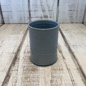 May include: A cylindrical ceramic tumbler with a smooth, matte finish in a muted slate blue color. The vessel features subtle horizontal ridges along its exterior wall and a simple, open top design, showcasing a consistent glaze throughout the piece.