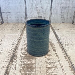 May include: A cylindrical ceramic tumbler featuring a deep blue and teal mottled glaze with subtle horizontal ridges along the exterior surface. The vessel has a smooth, dark interior and a straight, uniform profile, showcasing a handcrafted aesthetic.