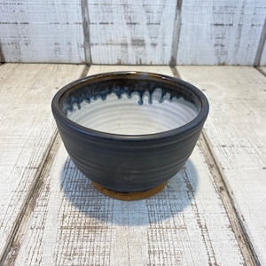 May include: A black ceramic bowl with a white glaze inside and a brown rim. The bowl has a slightly textured surface and is sitting on a wooden surface.