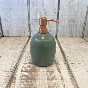 May include: A sage green ceramic soap dispenser with a smooth, rounded body and a metallic copper-colored pump mechanism. The dispenser features a speckled finish on the ceramic surface and a functional, downward-facing spout on the pump head.