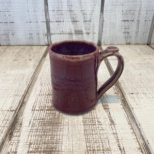 May include: A handmade ceramic mug in a deep burgundy color. The mug has a cylindrical shape with a rounded handle and a small thumb rest. The interior is a lighter shade of burgundy, and the exterior has subtle texture.