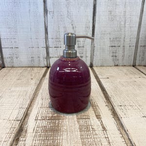 May include: A round, deep purple ceramic soap dispenser with a glossy, speckled finish. The container features a metallic silver-toned pump mechanism at the top, designed for dispensing liquid soap or lotion through a standard manual press action.
