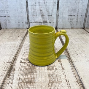 May include: A handmade, chartreuse-colored ceramic mug with a rounded handle. The mug has horizontal ridges around the body and a small, circular detail where the handle meets the mug. The mug sits on a distressed, white-painted wooden surface.