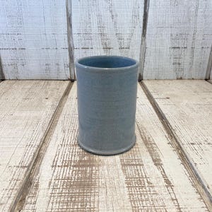 May include: A cylindrical ceramic tumbler with a smooth, matte finish in a muted slate blue color. The vessel features a simple, straight-walled design with a slightly flared base and a clean, open top rim, showcasing a uniform, solid color.