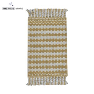May include: Rectangular rug with a repeating pattern of gold-colored blocks on a cream background. The rug has a fringe on both ends and the text "THE RUGS STORE" in the upper left corner.