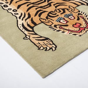 Op de afbeelding: Rechthoekig tapijt met een gestileerd tijgerontwerp. De tijger heeft een bruin lichaam met zwarte strepen, rode en zwarte accenten en blauwe ogen. Het tapijt heeft een lichtgroene achtergrond en is gemaakt van zacht materiaal.
