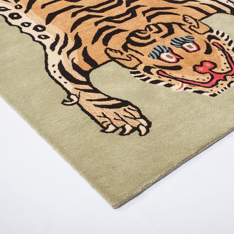 Op de afbeelding: Rechthoekig tapijt met een gestileerd tijgerontwerp. De tijger heeft een bruin lichaam met zwarte strepen, rode en zwarte accenten en blauwe ogen. Het tapijt heeft een lichtgroene achtergrond en is gemaakt van zacht materiaal.