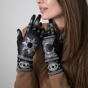 Op de afbeelding: Een vrouw draagt een bruine trui en zwarte handschoenen met zilveren borduurwerk van hemelse symbolen, waaronder een zon, een maan en ogen.