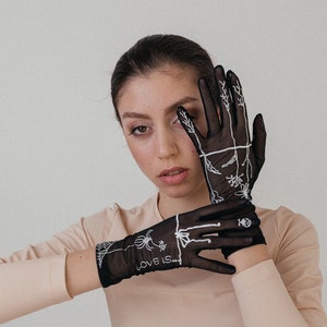 May include: A person wearing a pair of black sheer gloves with white embroidery. The gloves feature a spider web design and the text "LOVE IS..." on one glove.