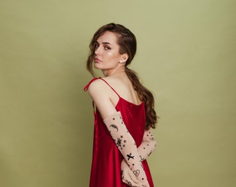 May include: A woman wearing a red satin dress with a pair of sheer gloves with black tattoo-like designs. The gloves are on her arms and hands. She is standing in front of a green background.