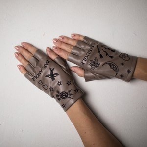 Könnte beinhalten: Ein Paar braune fingerlose Handschuhe mit schwarzer Stickerei. Ein Handschuh trägt den Text "LAVEUGLE" und der andere den Text "PAR AMOUR".