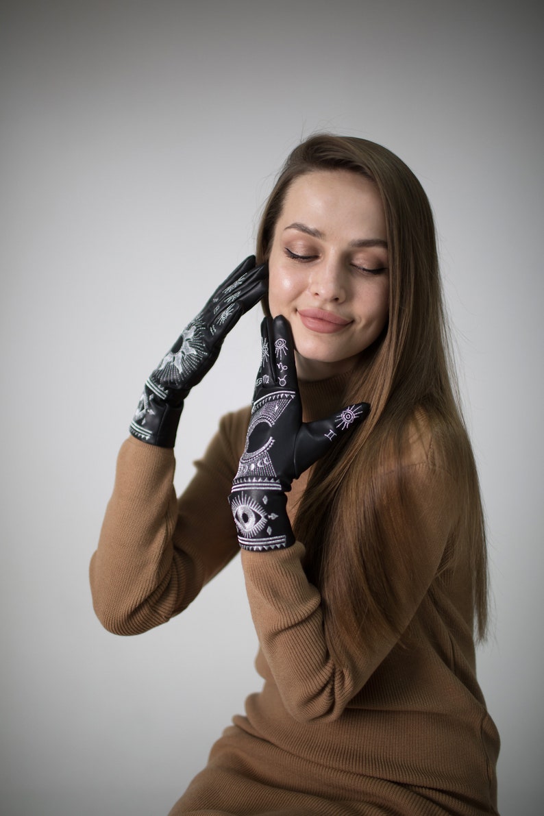 Op de afbeelding: Een vrouw draagt een bruine trui en zwarte handschoenen met witte borduursels. De handschoenen hebben een oogontwerp en andere symbolen.