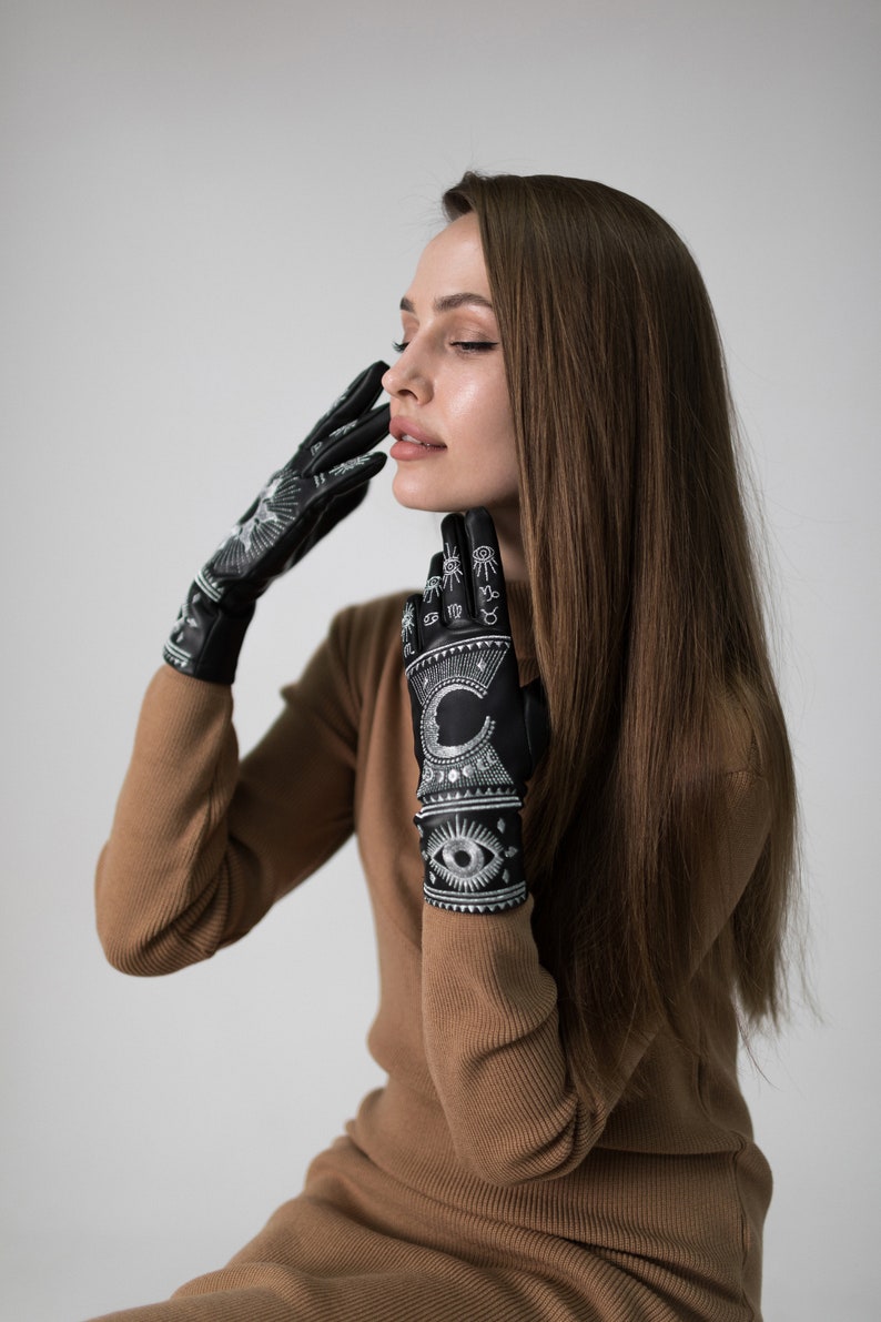 Op de afbeelding: Een vrouw draagt een bruine trui-jurk en zwarte handschoenen met witte astrologische symbolen en een oogontwerp.