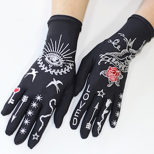 May include: A pair of black fingerless gloves with white embroidered designs, including a heart, a key, arrows, stars, a snake, birds, an eye, a rose, and the word "LOVED".