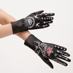May include: A pair of black leather gloves with white embroidered designs. The gloves feature various symbols, including a heart, stars, arrows, birds, and a snake. One glove has the word "Loved" embroidered on the back of the hand, and the other glove has the word "Forever" embroidered on the back of the hand.