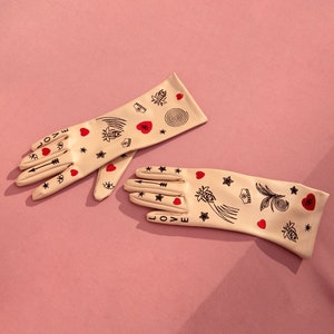 May include: A pair of beige gloves with a tattoo-inspired design. The gloves feature red hearts, stars, arrows, and other symbols. The word "LOVE" is written on the gloves.