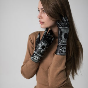 Op de afbeelding: Een vrouw draagt een bruine jurk en zwarte leren handschoenen met witte geborduurde zon-, maan- en oogontwerpen.