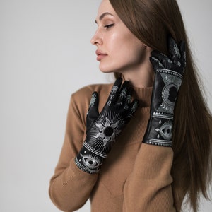 Op de afbeelding: Een vrouw draagt een bruine turtleneck trui en zwarte handschoenen met witte geborduurde ontwerpen. De handschoenen hebben zon-, maan- en oogmotieven.
