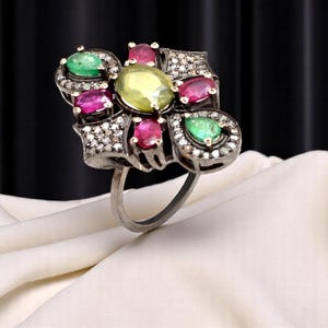 Op de afbeelding: Een zilveren ring met een cluster van kleurrijke edelstenen. De ring heeft een centrale ovale gele steen, omringd door roze en groene stenen en geaccentueerd met kleine heldere stenen. De ring ligt op een witte ondergrond.