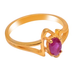 May include: A gold ring featuring an oval, deep red gemstone. The ring has a split shank design with a decorative setting around the gemstone. The ring is set against a white background.