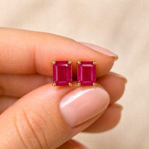 May include: A pair of gold stud earrings, each featuring a rectangular, vibrant red gemstone. The earrings are held between fingers, showcasing the polished gold setting and the rich color of the gemstones. The earrings are a classic jewelry item.