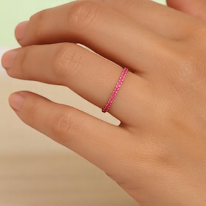May include: A bright pink braided ring is worn on a finger. The ring is a thin band with a textured, woven appearance. The ring's vibrant color contrasts with the skin tone, highlighting the jewelry.