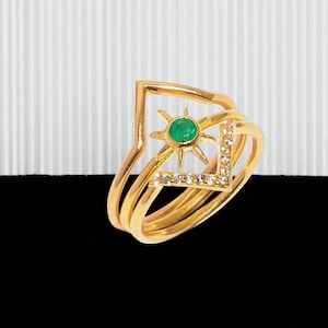 May include: A set of three gold rings. One ring features a V-shape design, another has a sunburst with a green center stone, and the third is a simple band. The sunburst ring has small, clear stones.